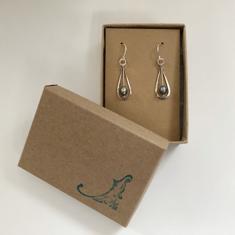 May include: A pair of silver dangle earrings with a dark gray pearl in a teardrop-shaped setting. The earrings are in a brown cardboard box.