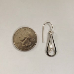 May include: A silver dangle earring with a small white pearl hanging from a teardrop-shaped frame. The earring is shown next to a US quarter for size comparison.