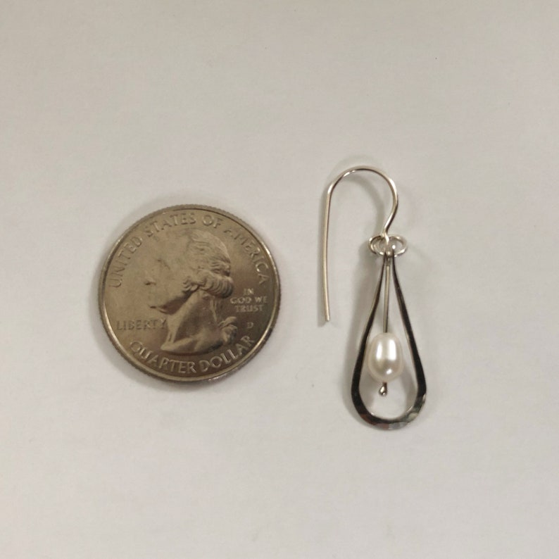 May include: A silver dangle earring with a small white pearl hanging from a teardrop-shaped frame. The earring is shown next to a US quarter for size comparison.