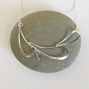 May include: A silver necklace with a hammered, abstract design. The necklace is made of multiple curved pieces that form a flowing, organic shape.