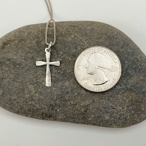 Puede incluir: Un collar con un colgante de cruz de plata en una cadena de plata. La cruz est&aacute; martillada y tiene un acabado texturizado. El collar se muestra en una roca junto a una moneda de 25 centavos de d&oacute;lar estadounidense para referencia de tama&ntilde;o.