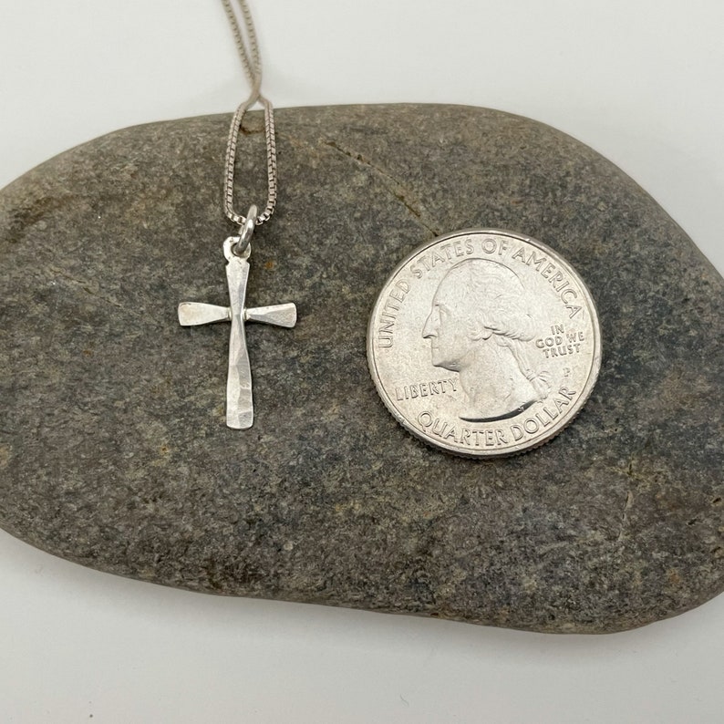 Puede incluir: Un collar con un colgante de cruz de plata en una cadena de plata. La cruz est&aacute; martillada y tiene un acabado texturizado. El collar se muestra en una roca junto a una moneda de 25 centavos de d&oacute;lar estadounidense para referencia de tama&ntilde;o.