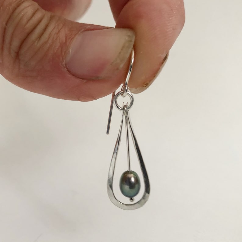 May include: Silver dangle earrings with a teardrop shaped design. A dark grey pearl hangs from the center of the teardrop.