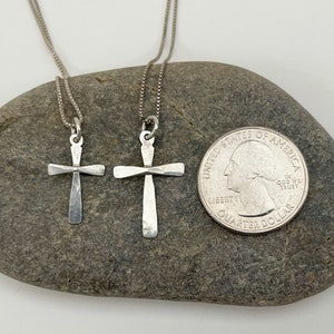 Puede incluir: Dos collares de plata con cruces sobre un fondo de roca gris. Las cruces son martilladas y tienen un aspecto r&uacute;stico. La cruz m&aacute;s grande es aproximadamente del tama&ntilde;o de una moneda de veinticinco centavos.