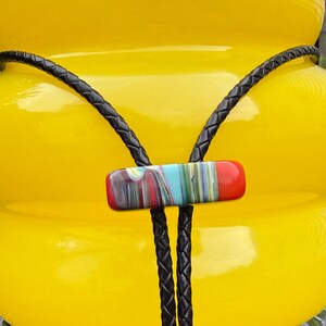 Op de afbeelding: Een heldergeel, bolvormig decoratief object met een zwart gevlochten koord en een rechthoekige bolo-stropdassluiting. De sluiting heeft een kleurrijk, gestreept ontwerp met rode, blauwe en groene tinten. Het object staat op een metalen oppervlak.