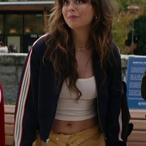 May include: A dark navy blue zip-up bomber jacket featuring long sleeves with white, red, and green vertical stripes along the outer arms, worn over a plain white cropped tank top paired with a tan-colored skirt with a layered waistband detail.