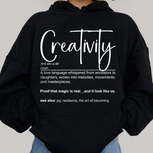 Może przedstawiać: Czarna bluza z kapturem z napisem "Creativity" w białej kursywie. Projekt zawiera definicję kreatywności i frazę "Proof that magic is real". Wygodny, codzienny element garderoby.