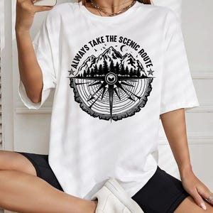 May include: White t-shirt with a black graphic featuring mountains, trees, a compass design, and the text "ALWAYS TAKE THE SCENIC ROUTE". The shirt has short sleeves and a crew neck.