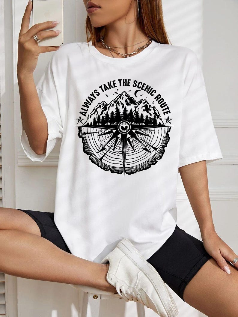 May include: White t-shirt with a black graphic featuring mountains, trees, a compass design, and the text "ALWAYS TAKE THE SCENIC ROUTE". The shirt has short sleeves and a crew neck.