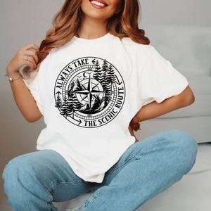May include: Cream-colored t-shirt featuring a black graphic of a compass rose, mountains, and trees. The text "ALWAYS TAKE THE SCENIC ROUTE" is printed around the compass. The shirt is a casual, comfortable style.