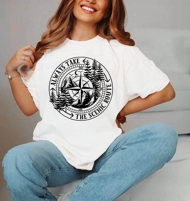 May include: Cream-colored t-shirt featuring a black graphic of a compass rose, mountains, and trees. The text "ALWAYS TAKE THE SCENIC ROUTE" is printed around the compass. The shirt is a casual, comfortable style.