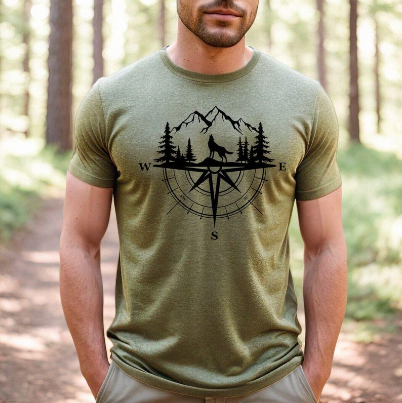 May include: Olive green t-shirt featuring a black graphic of a wolf howling at the moon, set against a mountain and compass rose design. The shirt has short sleeves and a crew neck.