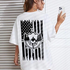 May include: White t-shirt with a black graphic on the back featuring an American flag design with a compass, mountains, and trees. The compass points North, South, East, and West. The shirt has short sleeves and a relaxed fit.