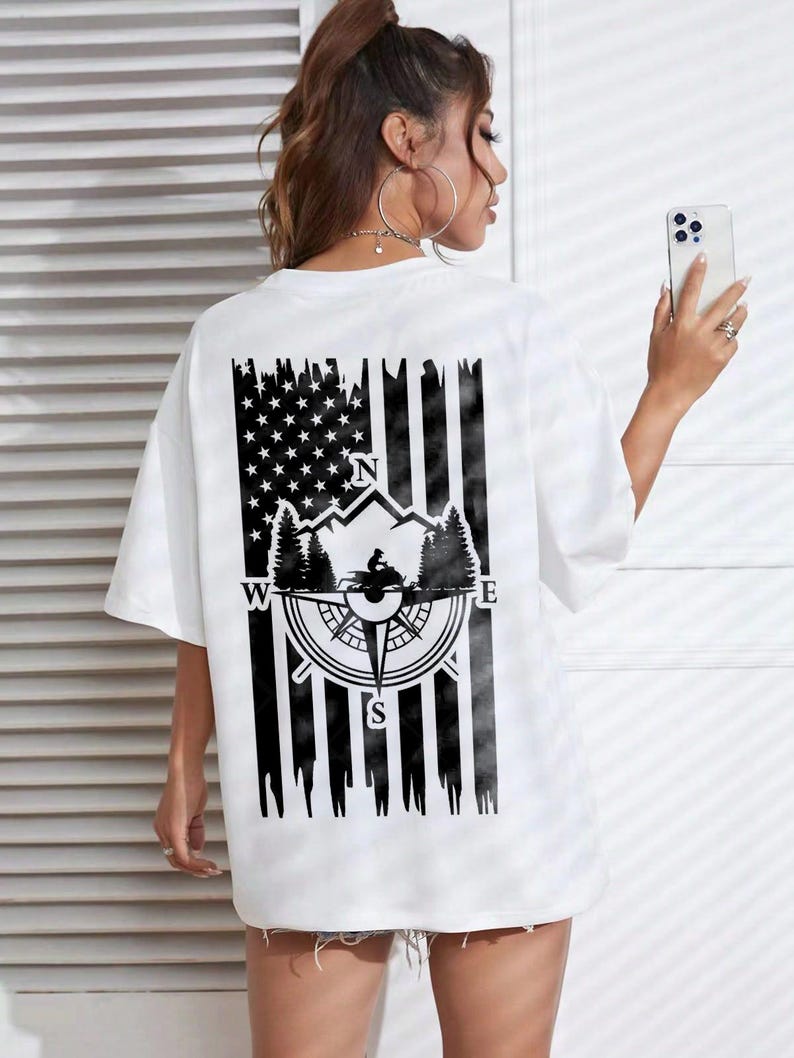 May include: White t-shirt with a black graphic on the back featuring an American flag design with a compass, mountains, and trees. The compass points North, South, East, and West. The shirt has short sleeves and a relaxed fit.