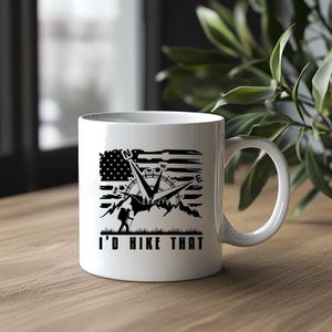 May include: White ceramic mug featuring a black graphic of a compass, mountains, and a hiker set against a distressed American flag design. The text "I'D HIKE THAT" is printed below the graphic. A perfect gift for hikers.