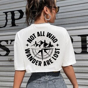 May include: White crop top with a black graphic design on the back. The design features a compass, mountains, and the text "NOT ALL WHO WANDER ARE LOST" in a circular arrangement. The model is wearing denim shorts.