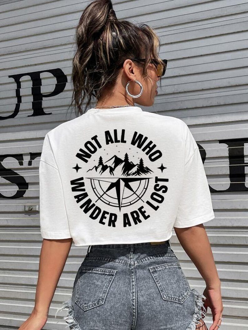 May include: White crop top with a black graphic design on the back. The design features a compass, mountains, and the text "NOT ALL WHO WANDER ARE LOST" in a circular arrangement. The model is wearing denim shorts.