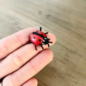 May include: A small, red ladybug figurine with black spots and legs. The ladybug has a black head and is resting on a finger. The figurine is a miniature representation of the insect.