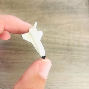 May include: A small, white toy space shuttle with black accents on the nose and wings. The miniature spacecraft is held between two fingers, showcasing its compact size and detailed design. The toy is likely made of wood or plastic.