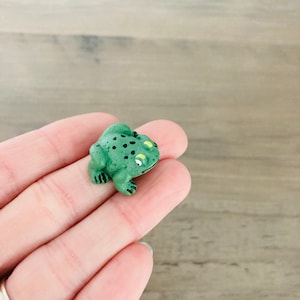 May include: A small green frog figurine with black spots and yellow eyes. The frog is sitting on a hand.