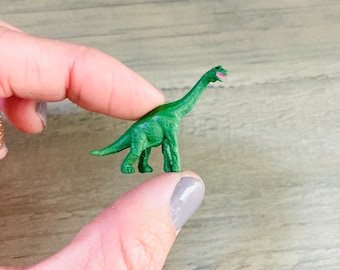 May include: A small green plastic dinosaur toy with a long neck. The dinosaur is standing on a person's finger.
