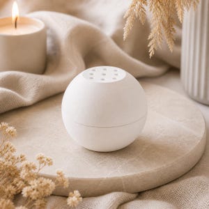 Könnte beinhalten: Weißer, kugelförmiger Diffusor für ätherische Öle mit kleinen Löchern oben. Der Diffusor steht auf einem runden, hellen Steintablett. Eine brennende Kerze und getrocknete Blumen im Hintergrund schaffen eine ruhige Atmosphäre. Der Diffusor hat einen Durchmesser von ca. 7 cm.