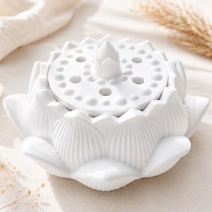 Könnte beinhalten: Ein weißer Räucherstäbchenhalter aus Keramik in Form einer blühenden Lotusblume. Der runde Deckel hat einen zentralen knospenförmigen Griff, der von mehreren kleinen kreisförmigen Löchern umgeben ist, und ruht auf einer Basis aus geformten Blütenblättern.