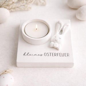 Könnte beinhalten: Ein weißer Kerzenhalter mit einer brennenden Teelichtkerze. Rechts ist ein weißes Häschen mit Fliege. Auf der Basis steht "kleines OSTERFEUER". Osterdekoration.