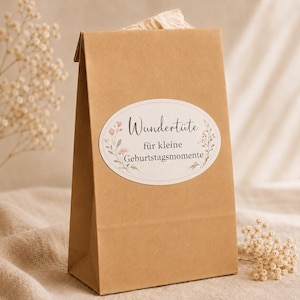 Könnte beinhalten: Eine braune Geschenktüte aus Kraftpapier mit einem ovalen Aufkleber, der florale Illustrationen und den Text Wundertüte für kleine Geburtstagsmomente zeigt. Die Tüte hat eine gefaltete Oberkante und besteht aus hellbraunem Papier.