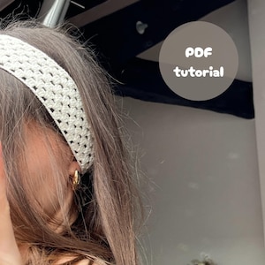 Peut inclure: Un bandeau en crochet blanc porté par une personne aux cheveux longs et bruns. Le bandeau est un modèle simple avec un motif répétitif. L'image comprend le texte "PDF tutorial".