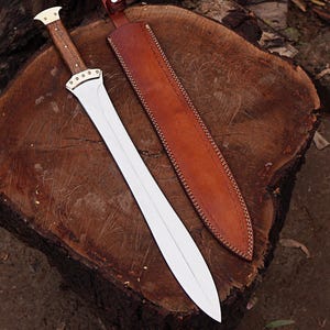 May include: A double-edged sword with a polished silver-toned metal blade, a wooden handle, and brass-coloured metal accents at the hilt. The sword is accompanied by a tan leather sheath featuring visible stitching and a small snap button closure.