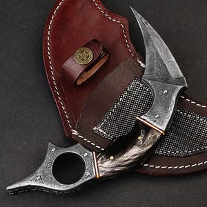 May include: A handcrafted, ornate knife with a curved blade and a dark, textured handle. The knife is displayed with a brown leather sheath, featuring a decorative button and white stitching. The blade has a unique pattern.