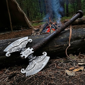 May include: A double-bitted battle axe featuring a silver-toned metal head with intricate etched patterns and a dark brown textured handle. The handle includes a braided grip section and a thin leather strap attached to the rounded pommel end.