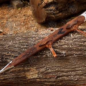 Puede incluir: Una vaina de espada de madera tallada con punta plateada y correa de cuero. La vaina presenta patrones grabados y un motivo de p&aacute;jaro. El fondo es una superficie de madera texturizada.