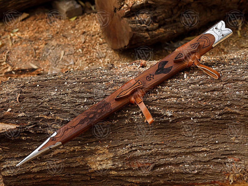Puede incluir: Una vaina de espada de madera tallada con punta plateada y correa de cuero. La vaina presenta patrones grabados y un motivo de p&aacute;jaro. El fondo es una superficie de madera texturizada.