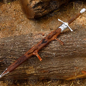 Puede incluir: Una espada artesanal con empu&ntilde;adura y vaina de madera marr&oacute;n, apoyada sobre un tronco de madera. La espada presenta una guarda plateada y un dise&ntilde;o detallado en la vaina. La hoja es plateada y puntiaguda.