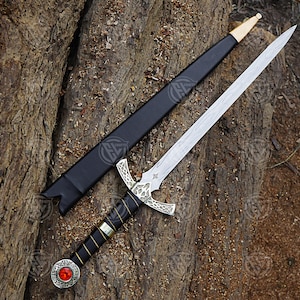 May include: An ornamental sword with a silver blade and decorative hilt, placed on a textured wooden surface. The hilt has a red gem and detailed silver patterns. A black sheath with gold accents is also present.