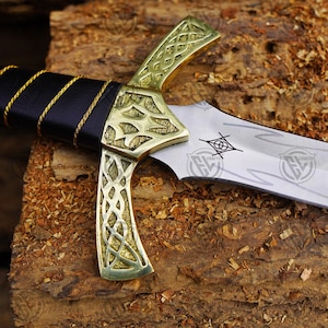 May include: A detailed view of a decorative sword. The blade is silver, and the hilt is gold-coloured with intricate patterns. The handle is wrapped in black leather with gold detailing. The blade has etched designs.