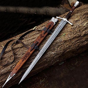 Puede incluir: Una espada y su vaina descansan sobre un tronco. La espada tiene una hoja plateada con un dise&ntilde;o y un mango marr&oacute;n. La vaina de madera presenta intrincadas tallas y correas de cuero.