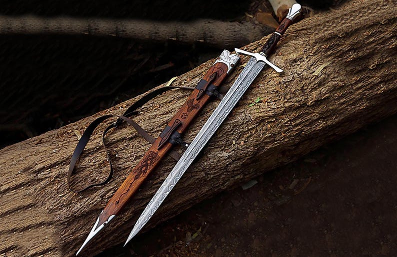 Puede incluir: Una espada y su vaina descansan sobre un tronco. La espada tiene una hoja plateada con un dise&ntilde;o y un mango marr&oacute;n. La vaina de madera presenta intrincadas tallas y correas de cuero.