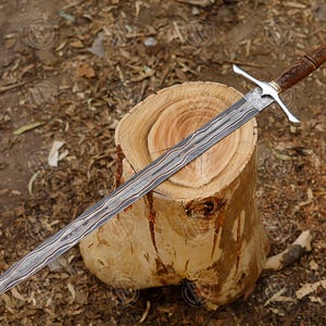 Puede incluir: Una espada larga y ornamentada con una hoja estampada descansa sobre un toc&oacute;n de madera. La empu&ntilde;adura es marr&oacute;n con intrincadas tallas y una guarda cruzada plateada. La hoja tiene un patr&oacute;n distintivo en forma de onda.