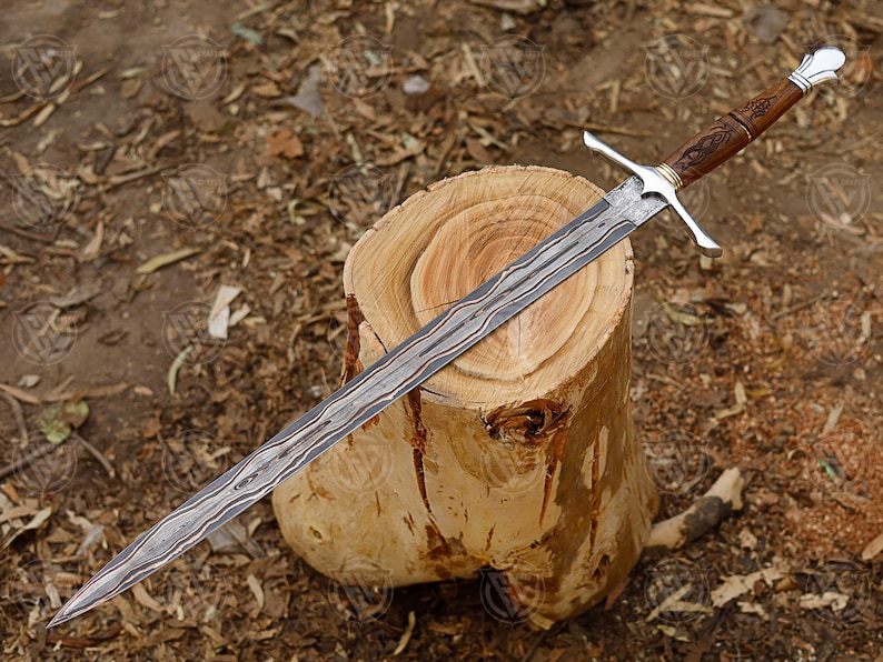 Puede incluir: Una espada larga y ornamentada con una hoja estampada descansa sobre un toc&oacute;n de madera. La empu&ntilde;adura es marr&oacute;n con intrincadas tallas y una guarda cruzada plateada. La hoja tiene un patr&oacute;n distintivo en forma de onda.