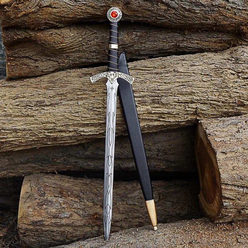 May include: A decorative sword with a silver blade and ornate hilt, featuring a red gem at the pommel. The sword has a black handle and is accompanied by a black sheath with a gold-coloured tip. The sword is leaning against a backdrop of wooden logs.