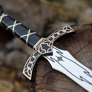 May include: A decorative sword with a black handle wrapped in gold-coloured thread. The hilt and blade feature gold and black accents. The pommel has a red gem. The sword rests on a wooden surface.