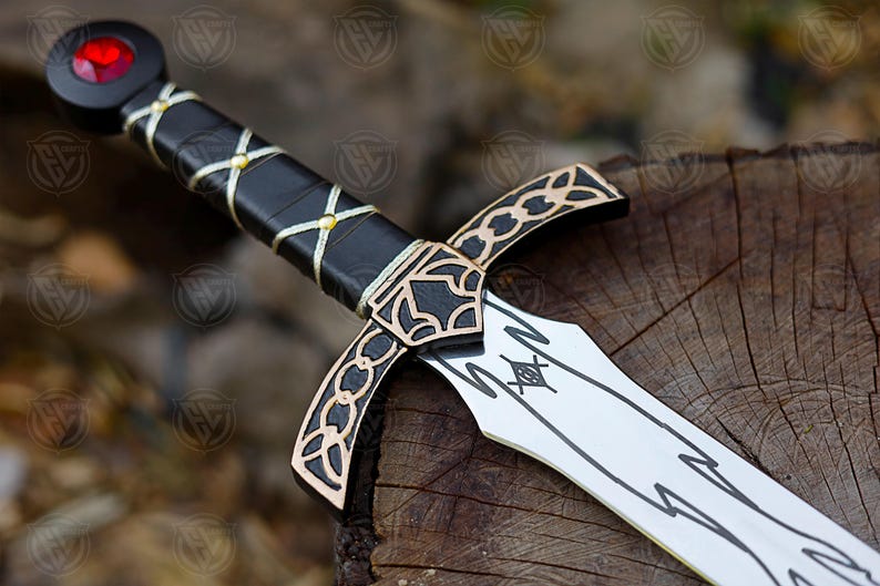 May include: A decorative sword with a black handle wrapped in gold-coloured thread. The hilt and blade feature gold and black accents. The pommel has a red gem. The sword rests on a wooden surface.