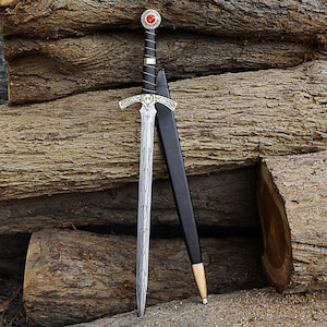 Puede incluir: Una espada de estilo medieval con una hoja plateada y una empuñadura ornamentada, con una gema roja en el pomo. La espada se combina con una vaina negra con detalles dorados. La espada se exhibe sobre un fondo de troncos de madera apilados.