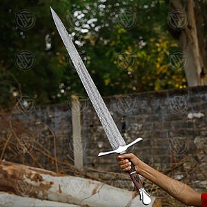 Puede incluir: Una espada larga y plateada con una hoja estampada y un mango marr&oacute;n y plateado. La espada se sostiene contra un fondo borroso de &aacute;rboles y una pared de ladrillos. La hoja mide aproximadamente 76 cm de largo.
