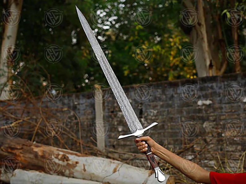 Puede incluir: Una espada larga y plateada con una hoja estampada y un mango marr&oacute;n y plateado. La espada se sostiene contra un fondo borroso de &aacute;rboles y una pared de ladrillos. La hoja mide aproximadamente 76 cm de largo.