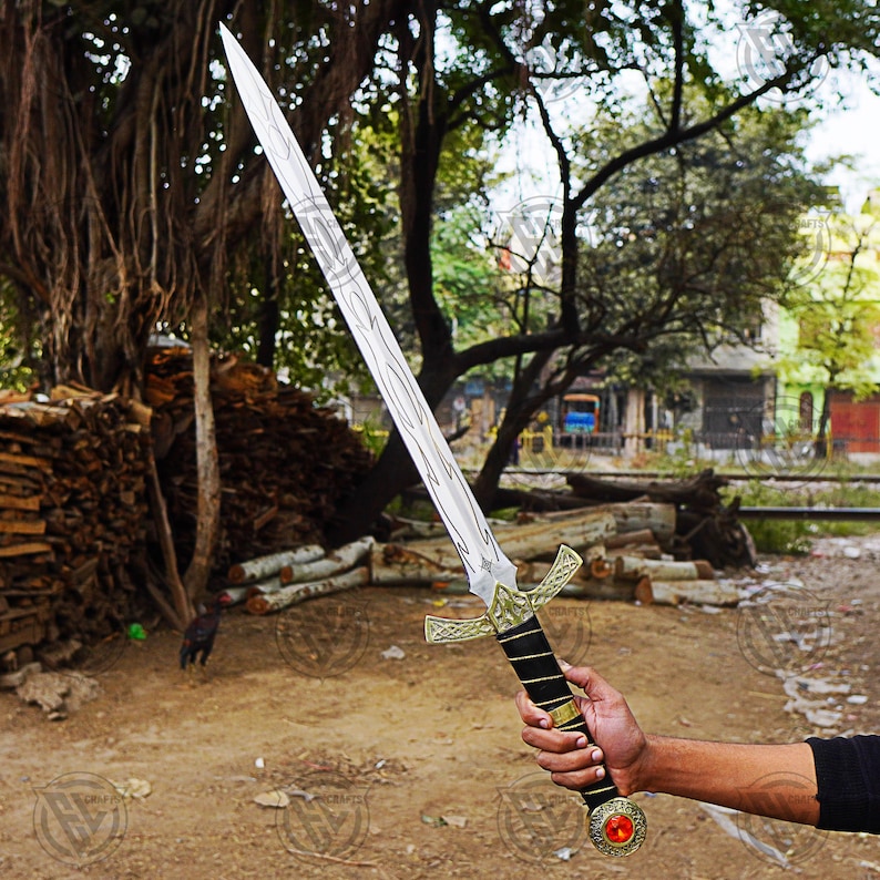 May include: A long, silver sword with a wooden-grain pattern on the blade and a gold and black handle. The sword has a crossguard with a Celtic knot design and a red gem at the pommel. The sword is held in a person's hand.