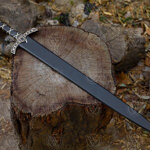 May include: A black sword with a gold and silver hilt, lying on a wooden surface. The sword has a red gem at the pommel and a decorative crossguard. The blade is black and the tip is gold.